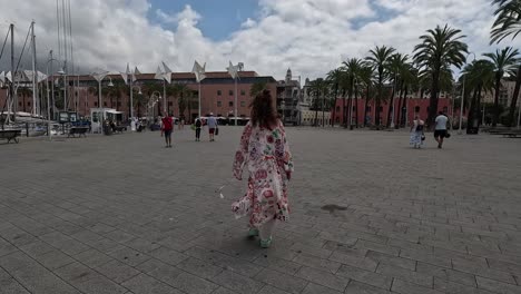 Following-shot-showing-lady-with-curly-hair-walking-through-town-square-in-Italy,-wearing-patterned-clothing