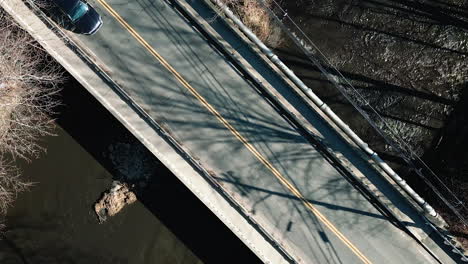 Drone-ascends-above-calm-empty-bridge-as-sedan-and-SUV-drive-across-asphalt,-long-tree-shadows
