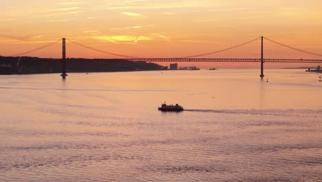 Tug-boat-ferry-crosses-Tagus-river-at-sunset-as-last-light-dusk-glow-spreads-on-silhouette-of-Suspension-bridge