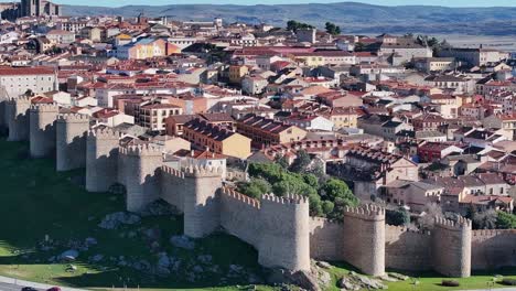70-mm-filming-with-a-drone-over-the-walled-city-of-Avila,-visualizing-the-roofs-of-the-houses,-the-stone-walls-and-their-towers,-a-road-with-vehicles-and-its-mountains-in-the-background