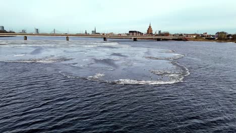 Luftaufnahme,-Der-Fluss-Daugava-Teilt-Die-Europäische-Stadt-Riga-In-Zwei-Hälften,-Vorn-Sieht-Man-Ein-Stück-Eis-Mit-Vielen-Möwen,-Die-Darauf-Treiben,-Verkehr-Auf-Der-Brücke
