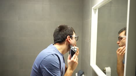 Side-closeup-shot-of-man-shaving-beard-with-an-electric-shaver,-mirror
