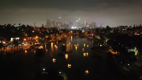 Glowing-swan-pedal-boats-on-Echo-Lake-with-the-Los-Angeles-city-skyline---aerial-flyover