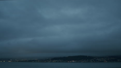 Heavy-dark-clouds-over-seaside-town-and-water,-dark-evening-with-distant-lights,-handheld