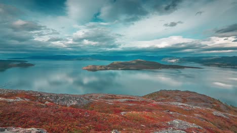 Stormy-clouds-backlit-by-the-autumn-sun-whirl-above-the-calm-fjord-and-mountains-in-the-timelapse-video