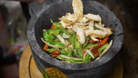 Cook-adds-sauted-peppers-onions-and-chicken-fajita-to-sizzling-hot-molcajete-lava-rock-bowl,-slow-motion-close-up-4K