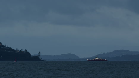 Distant-ferry-boat-on-the-water-coming-into-port-and-approaching-lighthouse-on-land-on-cloudy-day