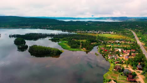 Aerial-Drone-View-of-a-Peaceful-Clear-Lake-with-Cars-Driving-next-to-a-Mountain-Town-with-Cabins-and-Vacation-Houses-in-a-Forest-with-Cloudy-Hills-in-the-Background