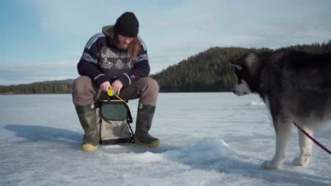Mann-Beim-Eisfischen-Mit-Seinem-Hund-Alaskan-Malamute-Auf-Dem-Zugefrorenen-See