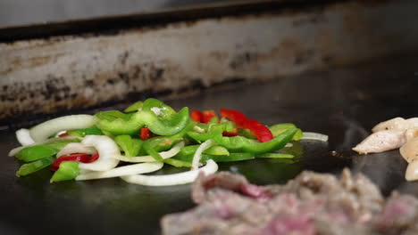 Gloved-hand-adds-sliced-red-green-bell-peppers-and-onions-to-steaming-hot-flat-top-grill-in-commercial-restaurant-kitchen-as-beef-chicken-fajitas-sizzle-in-foreground,-slow-motion-close-up-slider-4K