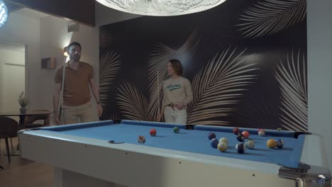 A-young-couple-is-playing-billiards,-the-billiards-mat-is-blue,-the-man-hits-the-cue-ball-with-his-cue-and-his-wife-watches-him