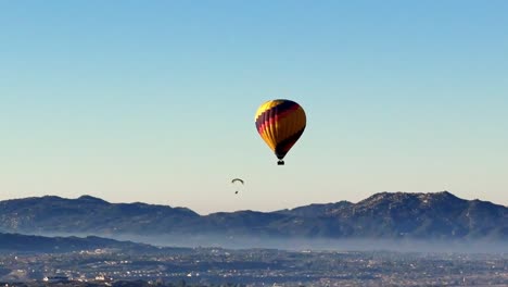Stationary-drone-view-of-a-Hot-Air-Balloon-and-a-Paraglider-sailing-by-from-right-to-left-over-Temecula-wineries-with-a-clear-sky-and-low-hanging-mist-and-hills-in-the-background