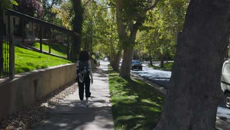 Woman-with-backpack-walks-on-sidewalk-shaded-by-trees,-Salt-Lake-City,-Utah,-USA