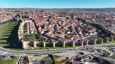 Vuelo-Con-Giro-De-Cámara-Visualizando-Con-Un-Cielo-Azul-La-Ciudad-De-ávila,-Patrimonio-Mundial-De-La-Unesco-Con-Su-Muro-De-Piedra-Y-Sus-Torres-Y-Una-Vía-Perimetral-Con-Vehículos-Circulando-En-España