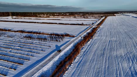 aerial-rural-car-scene-with-a-lone-vehicle-driving-on-a-snow-covered-road-that-winds-through-a-field,-with-a-line-of-dark-trees-in-the-distance-and-a-clear-blue-sky-overhead,-Orbit-Pan-Shot