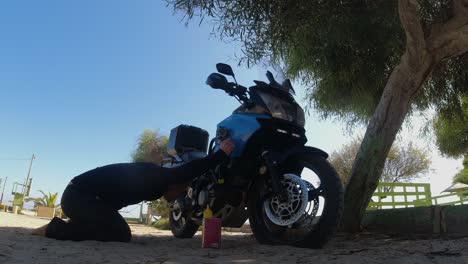 Young-man-checks-oil-level-of-motorcycle-in-shade-of-beach-tree