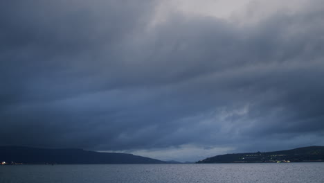 Schwere-Blaue-Wolken-Ziehen-Vorbei-Und-In-Der-Ferne-Rasen-Boote-Mit-Eingeschaltetem-Licht-Vorbei