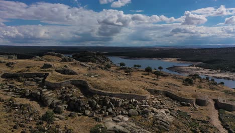 Vuelo-Lateral-En-Un-Fuerte-Celta-Vetton-De-La-Edad-De-Hierro-Viendo-La-Entrada-Al-Pueblo-Y-Su-Extenso-Muro-De-Piedra-Reconstruido-Al-Final-Vemos-La-Presa-Del-Embalse-En-Avila-España