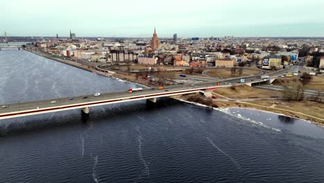 Aerial-of-highway-bridge-spanning-a-wide,-blue-river-Daugava-in-a-densely-populated-city-Riga-with-tall-skyscrapers-in-the-background,-Aerial-Orbit-Pan-Shot