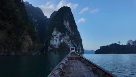 Front-of-traditional-wooden-boat-cruising-over-calm-water-in-Thailand-moving-towards-limestone-rock-formations-and-mountains-with-dense-native-rainforest