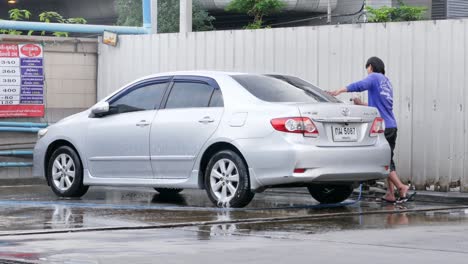 Rinsing-off-the-soapy-suds-off-a-car-using-a-sprinkler-attached-to-hose,-a-carwash-worker-is-cleaning-the-sides-and-under-chassis-of-a-vehicle-in-a-shop-in-Bangkok,-Thailand