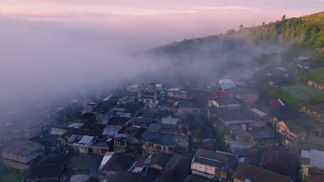 Mist-flowing-over-small-town-during-bright-sunset-of-sunrise,-aerial-view