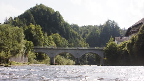 Historische-Steinbrücke-über-Einen-Malerischen-Fluss-In-Der-Sonne,-Berglandschaft