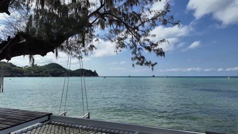 Luxury-hotel-decking-with-hammock-and-wooden-swing-at-the-shoreline-of-tropical-island-paradise,-with-views-to-horizon-of-calm-clear-ocean-water