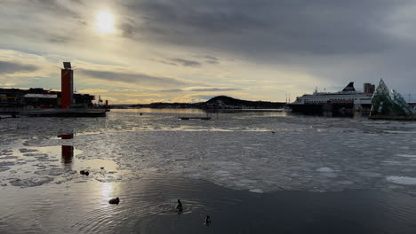 Oslo,-Norwegen-–-Gefrorener-Fjord-Mit-Schwimmenden-Enten-Und-Einem-In-Der-Ferne-Angedockten-Schiff-Bei-Sonnenuntergang-–-Weitwinkelaufnahme