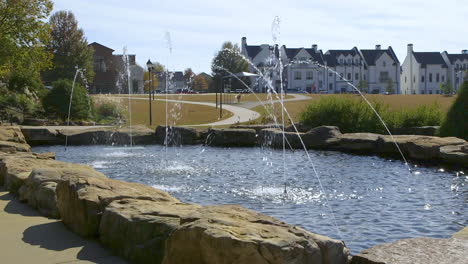 Public-park-pond-area-with-several-fountains-spraying-streams-of-clear-water-with-homes-and-buildings-in-the-background,-on-a-sunny-day