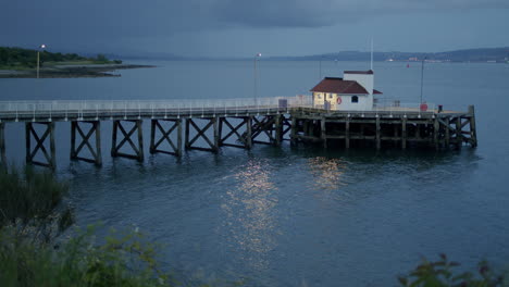 Old-wooden-pier-in-evening-light,-with-water-and-distant-flashing-lights,-static-wide-with-vegetation-around