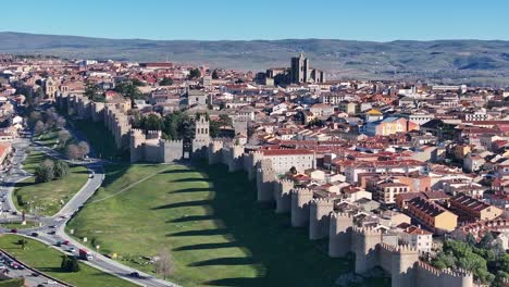 flight-in-the-northern-canvas-of-the-walled-city-of-Avila,-seeing-inside-the-cathedral-and-its-homes,-we-see-the-shadow-of-the-wall-projected-on-the-grass-and-a-road-with-cars-circulating-in-Spain