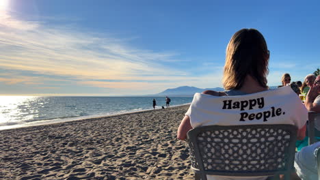 Woman-sitting,-talking-and-enjoying-life-at-a-beach-restaurant-in-Marbella,-woman-wearing-a-pullover-with-happy-people-words,-sunny-summer-day-in-Spain,-4K-shot