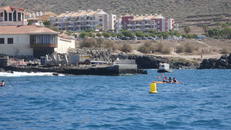 Tourists-kayaking-in-the-sea-near-a-rocky-shore,-with-urban-buildings-in-the-distance