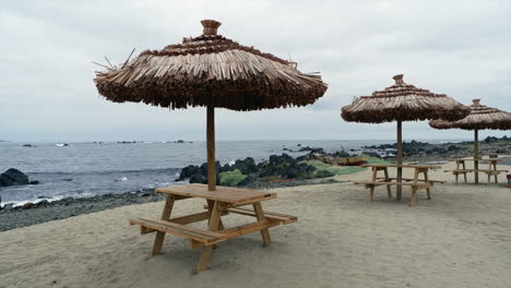 Overcast-beach:-Grass-umbrellas-over-empty-picnic-tables,-no-people