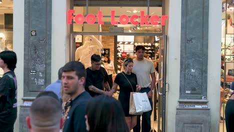 Customers-are-seen-shopping-at-the-American-multinational-sportswear-and-footwear-retailer,-Foot-Locker,-store-as-pedestrians-walk-past-the-frame-at-a-busy-retail-street