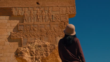 Asian-tourist-woman-visiting-Egypt-standing-in-front-of-Egyptian-hieroglyphs-carved-red-wall-in-Ancient-Egypt,-Egyptian-lost-language-in-oasis-North-Africa-desert