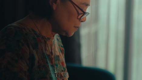 asiatic-senior-woman-wearing-glasses-sitting-alone-in-her-house-crafting-crochet-wool-while-looking-out-of-the-windows