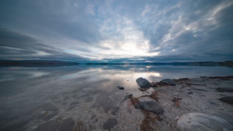 Storm-clouds-move-quickly-above-the-calm-fjord-and-pebble-beach,-captured-in-a-timelapse-video