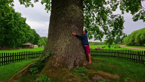 Young-man-hugging-massive-oak-tree-in-countryside-landscape,-dolly-backward