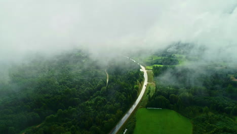Vista-Aérea-De-La-Carretera-Rural-De-Letonia-Que-Atraviesa-El-Bosque-Rodeada-De-Nubes.