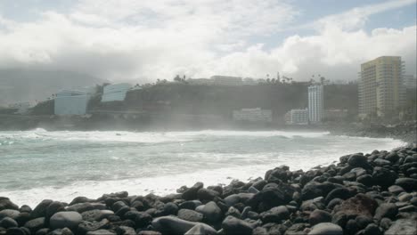 Slowmotion-surfers-waiting-on-ocean-wave,-pebble-beach-near-hotel-resorts,-Playa-Martiánez,-Puerto-de-la-Cruz,-Canary-Islands-in-spring