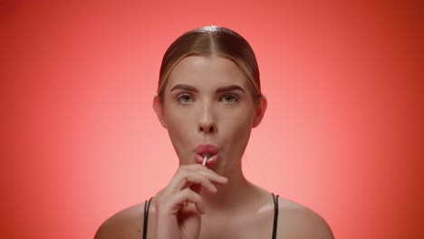 Childish-young-woman-licks-a-lollipop-and-smiling-at-camera,-studio-shot