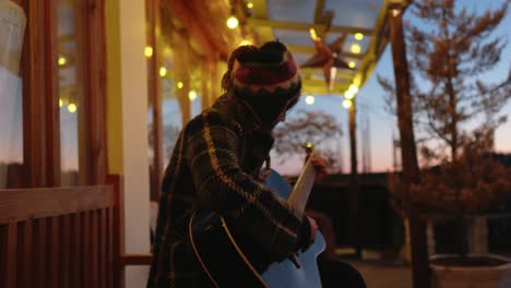 Side-view-of-a-woman-playing-guitar-on-a-porch-at-twilight-with-string-lights