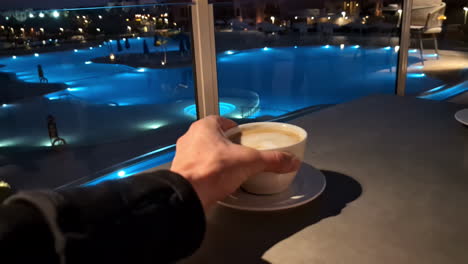 POV-of-hand-picking-up-coffee-from-table-with-luxurious-swimming-pool-backdrop