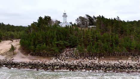 Aerial-white-lighthouse-on-a-hill-overlooking-a-body-of-water,-surrounded-by-green-trees,-cloudy-day,-Right-Truck,-Uzava-Lighthouse