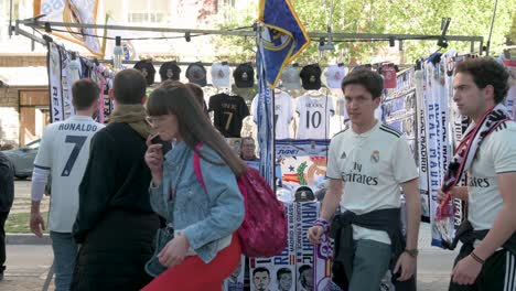 Un-Puesto-Callejero-Que-Vende-Productos-Del-Real-Madrid,-Como-Bufandas-Y-Camisetas,-Mientras-Los-Aficionados-Pasan-Junto-Al-Marco-Antes-De-Asistir-Al-Partido-De-Fútbol-De-La-Liga-De-Campeones-Contra-El-Equipo-De-Fútbol-De-Manchester-City.