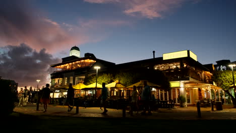 Visitors-walk-in-front-of-lit-up-restaurants-of-Hermanus-Waterfront,-twilight