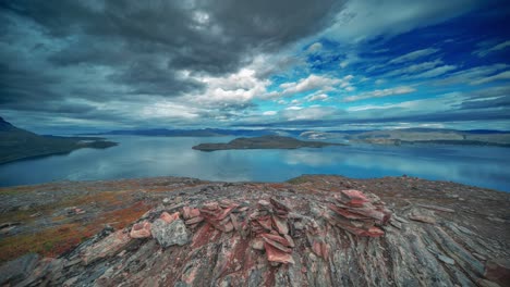 Whirling-stormy-clouds-are-backlit-by-the-setting-sun-above-the-serene-fjord-and-withered-mountains-in-the-time-lapse-video