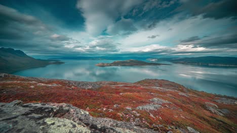 Whirling-stormy-clouds-are-backlit-by-the-low-sun-over-a-calm-mirror-like-fjord-and-autumn-tundra-in-the-timelapse-video